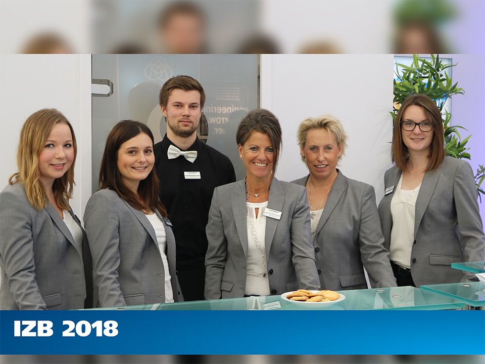 Fünf Personen in Geschäftskleidung stehen hinter einem Empfangstresen mit einem Teller voller Kekse; unten ist der Schriftzug IZB 2018 zu sehen.
