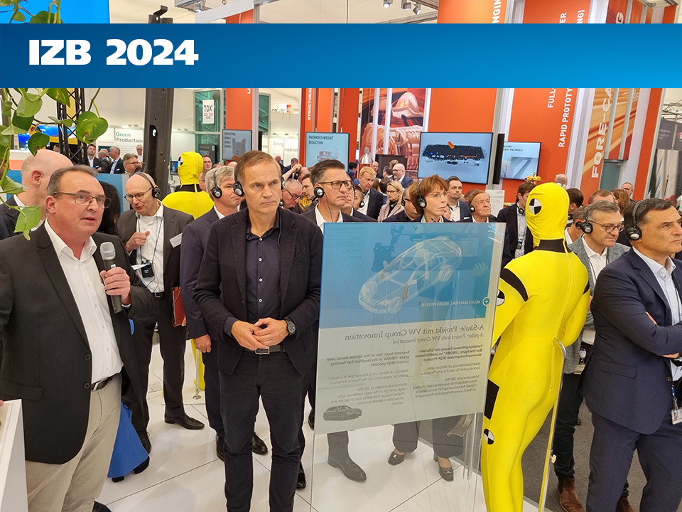 Ein Mann spricht vor Publikum in ein Mikrofon an einem Messestand, an dem ein gelber Crashtest-Dummy und ein Fachplakat zum Thema Automobilbau ausgestellt sind.